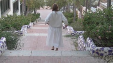 Beyaz elbiseli bir kadın zarif bir şekilde yeşil çalı ve pırıl pırıl bir heykel ile kaplı bir merdivenden iniyor, ağaçlı bir kaldırım ve su özelliğinin zeminine kurulmuş.