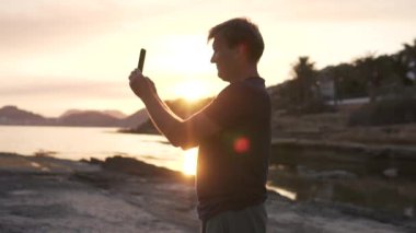 Bir adam gün batımında bir cep telefonuyla denizin fotoğrafını çekiyor. İspanya, Cabo de Huertas
