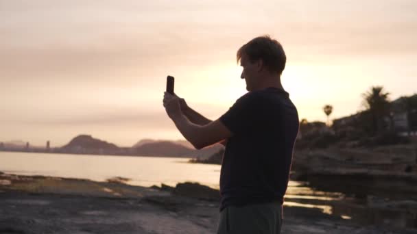 Przystojny Mężczyzna Robi Panoramiczne Zdjęcie Telefonie Kamieniami Morzem Zachodzącym Słońcu — Wideo stockowe