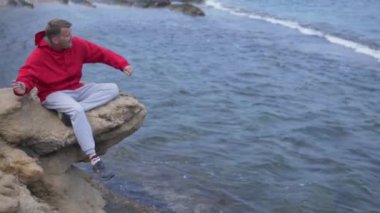 Şortlu bir adam sahildeki su kıyısındaki bir kayada oturur, rüzgar dalgalarının tadını çıkarır ve kıyıda dengede durur, okyanusa bakarken eğlence ve eğlenceye katılır.