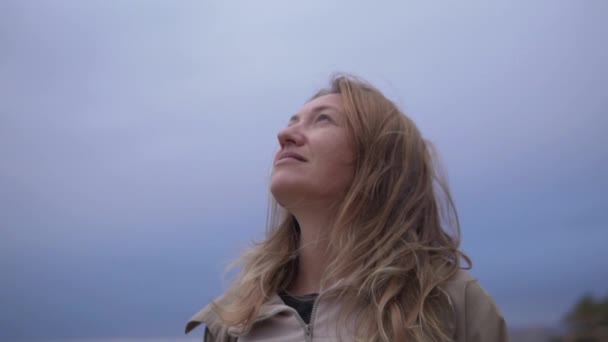 一个金发碧眼的女人站在一个充满活力的蓝天前 带着快乐的表情凝视着天空 她穿着一件风衣 这给影片情节增添了神秘感 — 图库视频影像