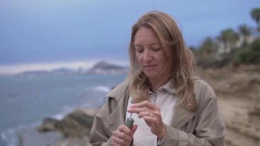 Okyanus kıyısındaki kayalık bir sahilde duran bir kadın, elektronik sigaraya bir sigara yerleştirir, sörfçü saçlarında rüzgarı hisseder ve suyun ve gökyüzünün güzel manzarasına hayran kalır.