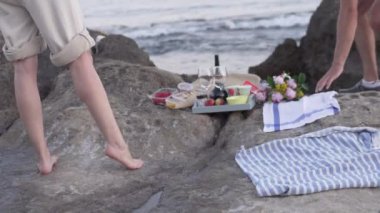 Romantik bir plaj pikniği yapan, okyanusun kenarında diz çöken ve suyun kenarında yan yana duran bir çift. Kadın bir tepsi yiyecekle rahatlar ve manzaranın tadını çıkarır.