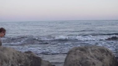 Plajda bir kadının okyanusa doğru yürüdüğü, bir kayanın üzerinde oturduğu ve kayalık bir sahilde durduğu video kaset. Rahatlatıcı, su ve kayaların yakın planlarını gösterir.