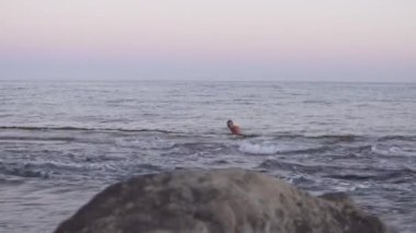 Bir adam okyanusta bir kayanın yanında yüzer, sonra da sahile doğru yürür. Video, deniz kenarında büyük bir kayayı gösteriyor. Kıyı şeridini manzaralı bir şekilde gösteriyor.
