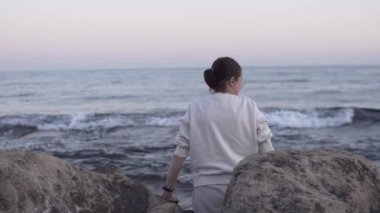 Okyanusun kenarında bir kadının durduğu, eğildiği ve bir kayanın üzerinde oturduğu, manzaranın tadını çıkardığı sakin bir video. Rahatlama veya doğal temalı projeler için uygundur