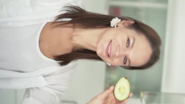 Dikey video. Beslenme uzmanı. Beyaz gömlekli sağlıklı genç bir kadının yakın plan görüntüsü ve parlak odanın bulanık arka planında gözünün üstüne avokado parçası tutması. Mutlu bayan doğal güzelliğe ulaşıyor.
