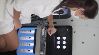 10 Ekim 2024, İspanya, Alicante, San Juan Güzellik Salonu. Tıbbi maskeli bir kadın kozmetik uzmanı çok işlevli cilt bakım cihazını çalıştırıyor. Dikey video