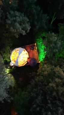 Huzurlu bir ormanda gece vakti bir hava yolculuğu, karanlık gökyüzüne karşı aydınlanan büyüleyici yapılar, bir kubbe çadırı ve bir ağaç ev gibi.