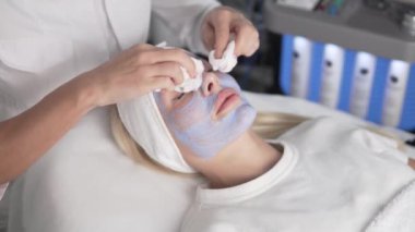 Kozmetik uzmanı müşterilerin yüzündeki mavi kil maskeyi çıkarıyor. Yakın plan.