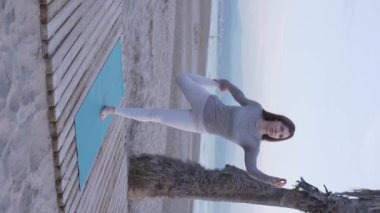 Güzel esmer kız sabahları deniz kıyısında yoga yapıyor ve esneme antrenmanı yapıyor. Dikey video