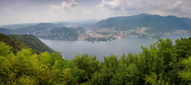 Como Gölü 'nün yemyeşil tepeler ve uzak kasabalarla çevrili nefes kesici panoramik manzarası. Sakin sahne, İtalya 'daki doğal güzellik ve sükunetin özünü yansıtıyor..