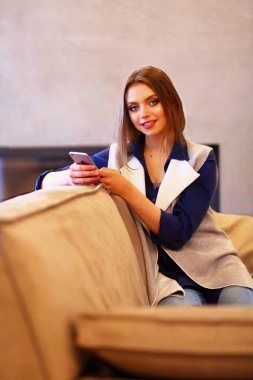 Genç bir kadın, beyaz odada rahat bir koltukta, elinde kahveyle oturuyor..