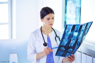 Doktor muayenehanesinde steteskoplu gülümseyen genç bayan doktor röntgeni tutuyor.
