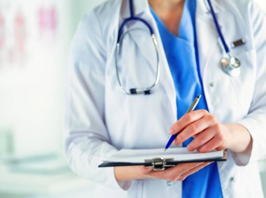 Steteskoplu bir doktor elinde bir defter tutuyor. Doktor elinde stetoskopla hastanede dikilirken tıbbi form dolduran kadın bir doktorun yakın çekimi.