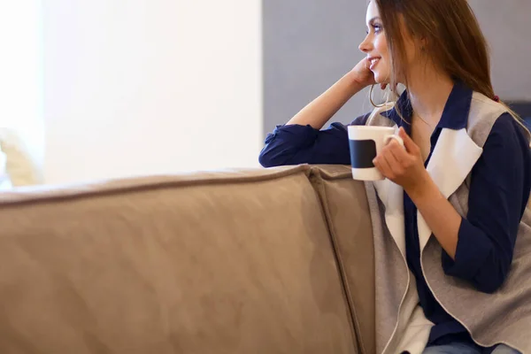 年轻女子坐在舒适的沙发上 手里拿着一杯咖啡 躺在白色的房间里 — 图库照片