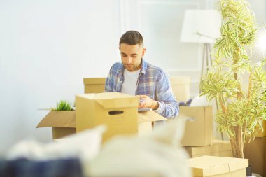 Taşınan bir adam boş bir dairede oturuyor, Kutuların arasında, eşyaların listesini kontrol ediyor.