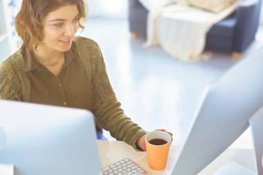 Çekici, gülümseyen bir kadın büro masasında oturuyor, elinde bir fincan kahve tutuyor, rahatlıyor ve başka tarafa bakıyor..
