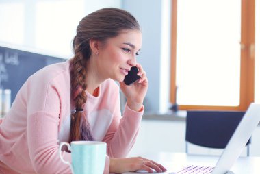 Genç kadın bir dizüstü bilgisayar kullanarak ve cep telefonuyla konuşurken mutfak masasında oturur