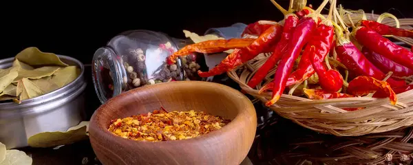 Wooden bowl of chili flakes, spices, bay leaf, red chili peppers still-life on black background with reflection