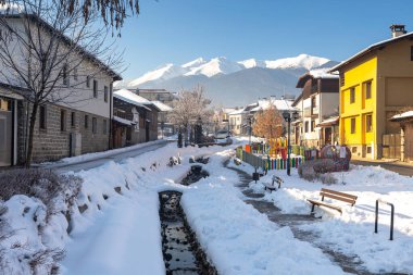 Bansko, Bulgaria - January 18, 2025: Gotse Delchev street view with old, traditional houses, water stream and Pirin mountains in winter clipart