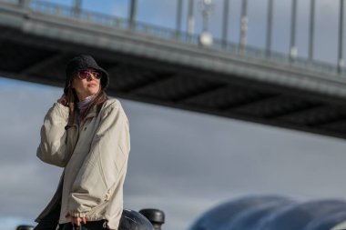 Sonbahar kadını sonbahar şehrinde geziniyor. Arka planda köprü ve gökyüzü olan günlük kıyafetler giyen güzel bir kadın.