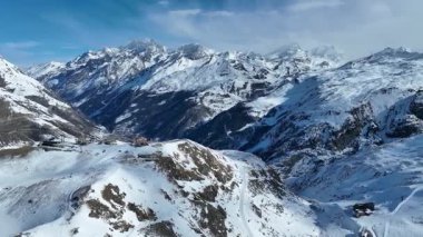 Gökyüzü arka planında yüksek karlı dağlar. Drone görüntülü teleferik istasyonu ve karlı dağ zirvesinde ulaşım için kablo yolu. Kış sırtı ve yamaçları