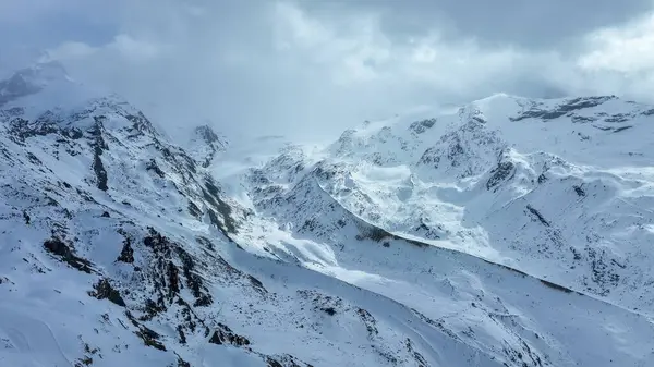 İsviçre 'nin Zermatt kentindeki karlı dağ zirvesinde kayak kaldırma kabloları, kayalık çıkıntılar ve bulutlu gökyüzünün altında bir zirve yapısı yer alıyor..
