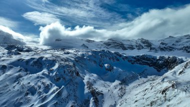 Yeşil kulelerde kar kaldıran gondollarla kaplı karlı dağ manzarası, arka planda engebeli zirveler, ve kısmen çıplak toprakla kaplı yamaçlar..