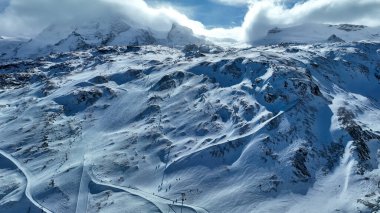 Beyaz manzaraya dağılmış kayakçılar ve sandalyeli geniş karlı kayak yamaçları. Etrafı engebeli dağlarla ve bulutlu gökyüzüyle çevrili..