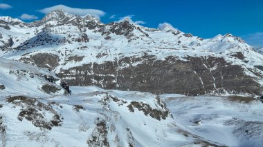 İsviçre Alpleri 'ndeki karla kaplı dağ zirveleri açık mavi gökyüzünün altında doğal güzellikleri ve dramatik dağlık manzaraları sergiliyor..