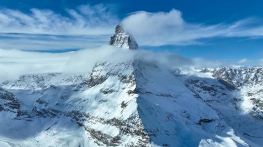 Bulutlarda gizlenmiş ikonik Matterhorn zirvesi karlı dağlık arazinin üzerinde görkemli bir şekilde yükselir. Çarpıcı kış manzarası, İsviçre Alplerinin parlak mavi gökyüzünün altındaki dokunulmamış güzelliğini gözler önüne seriyor..