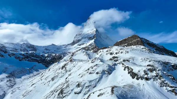 Bulutlarda gizlenmiş ikonik Matterhorn zirvesi karlı dağlık arazinin üzerinde görkemli bir şekilde yükselir. Çarpıcı kış manzarası, İsviçre Alplerinin parlak mavi gökyüzünün altındaki dokunulmamış güzelliğini gözler önüne seriyor..