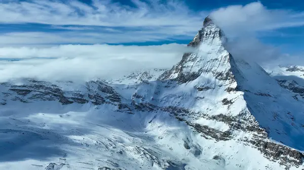 Görkemli Matterhorn, kısmen yumuşak bulutlarla örtülü, canlı mavi bir gökyüzünün altında dimdik ayakta duruyor. El değmemiş kar tarlaları, İsviçre Alpleri 'nin kışın nefes kesici güzelliğini vurgular..