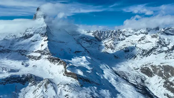 Görkemli Matterhorn, kısmen yumuşak bulutlarla örtülü, canlı mavi bir gökyüzünün altında dimdik ayakta duruyor. El değmemiş kar tarlaları, İsviçre Alpleri 'nin kışın nefes kesici güzelliğini vurgular..