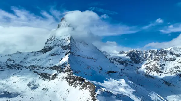 Görkemli Matterhorn, kısmen yumuşak bulutlarla örtülü, canlı mavi bir gökyüzünün altında dimdik ayakta duruyor. El değmemiş kar tarlaları, İsviçre Alpleri 'nin kışın nefes kesici güzelliğini vurgular..