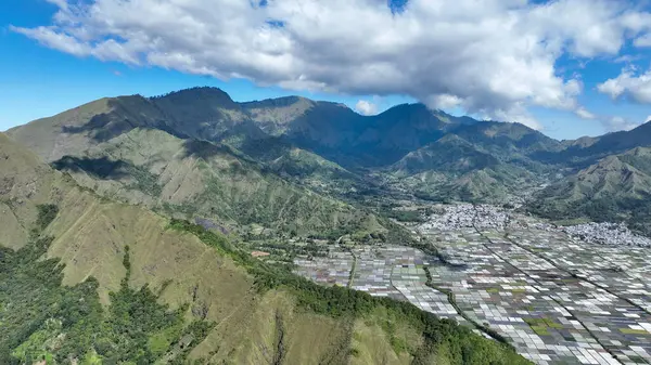 Arkasında yemyeşil dağlar olan Sembalun Vadisi 'nin panoramik hava manzarası parçalı bulutlu bir gökyüzünün altındaki renkli tarım alanlarıyla çelişiyor..