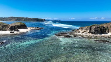 Kristal berrak turkuaz su dramatik kayalık kayalıklar ve beyaz kumla buluşur, dalgalar parlak mavi gökyüzünün altındaki uzak burunlara doğru uzanır..