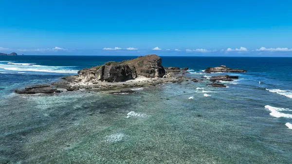 Engebeli bir kaya oluşumu kıyı şeridine hükmediyor. Sığ resifler ve canlı mavi sularla çevrili. Çarpıcı bir tropik deniz manzarası yaratıyor..