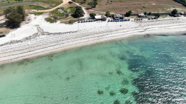 Lombok 'ta temiz turkuaz suyu olan el değmemiş beyaz bir kumsalın yakın çekimi. Plaj şemsiyeleri ve küçük kulübeler rahatlatıcı bir tropikal kaçış için davet ediliyor..