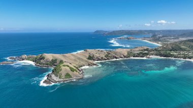 Derin mavi deniz ve turkuaz sığ sularla çevrili engebeli bir Lombok yarımadasının hava manzarası. Dalgalar kayalık kayalıklara çarparken yeşil tepeler okyanus kıyısında buluşuyor..
