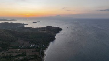 Alacakaranlıkta Lombok 'un hava görüntüsü engebeli kıyı şeridini ve dağınık adaları gösteriyor, solan güneşle gökyüzünü ve denizi yumuşak turuncu ve mor tonlarda boyuyor..