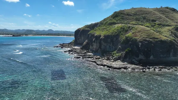 Dik yeşil tepeler kayalık bir kıyı şeridinin üzerinde yükselir, nazik dalgaların kıyıya vurduğu ve denizin sonsuz mavi tonlarda parıldadığı yerde