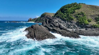 Görüntü, okyanus dalgalarının kayalık bir kıyı şeridine çarptığını gösteriyor. Dalgalar canlı bir turkuazdır, kayaların daha karanlık ve dünyevi tonlarıyla çelişir..