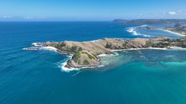 Derin mavi deniz ve turkuaz sığ sularla çevrili engebeli bir Lombok yarımadasının hava manzarası. Dalgalar kayalık kayalıklara çarparken yeşil tepeler okyanus kıyısında buluşuyor..