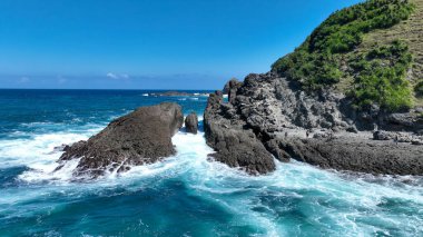 Görüntü, okyanus dalgalarının kayalık bir kıyı şeridine çarptığını gösteriyor. Dalgalar canlı bir turkuazdır, kayaların daha karanlık ve dünyevi tonlarıyla çelişir..