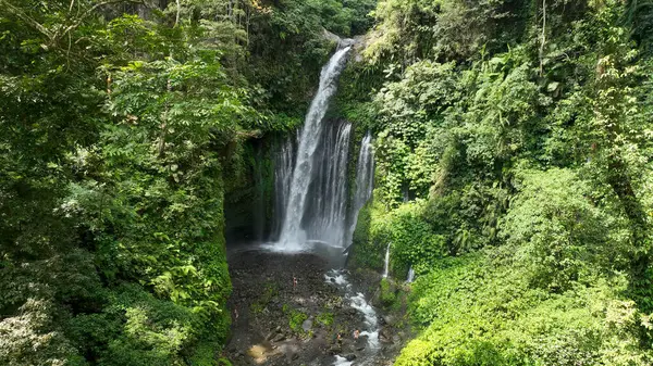 Tiu Kelep şelalesinin nefes kesen bir görüntüsü kayalık bir havzaya düşerken tüm yüksekliğini gözler önüne seriyor ve aşağıdaki küçük ziyaretçiler bu doğal harikanın ölçeğini vurguluyor..