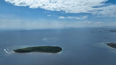 Geniş bir tropikal gökyüzü altında, yumuşak dalgalar ve diğer adaların uzak siluetleriyle çevrili, engin mavi denizde küçük yeşil bir ada yüzer..
