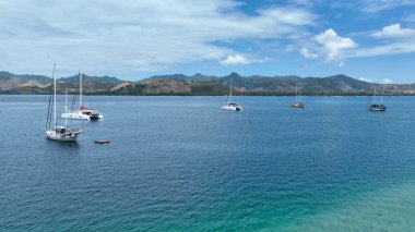 Yelkenliler ve katamaranlar arka planda yuvarlanan yeşil tepelerle derin mavi sularda huzur içinde yüzerler. Sahne, açık denizde sükunet ve macera saçar..