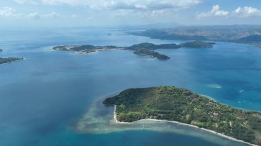 Bereketli bir tropikal adanın havadan görünüşü kristal berrak turkuaz sularla ve parlak mercan resifleriyle çevrili, ufuktaki uzak tepeler ve dağınık bulutlarla..